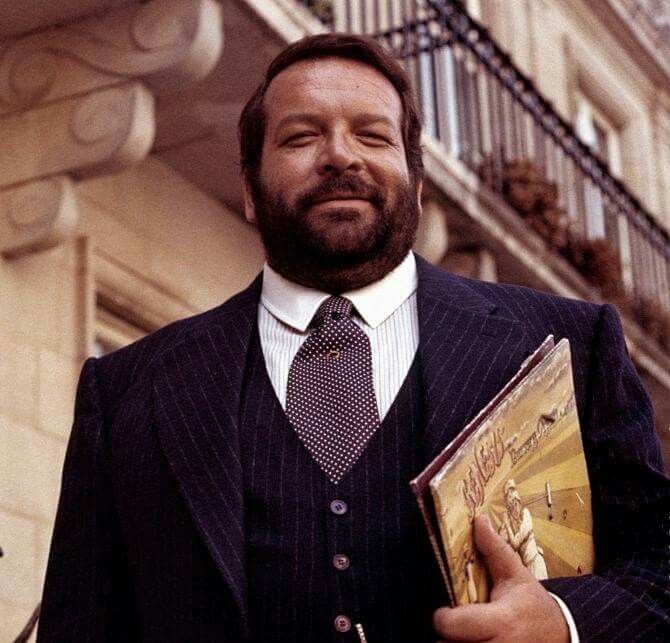 a man wearing a suit and tie holding a book in his hand while standing next to a building