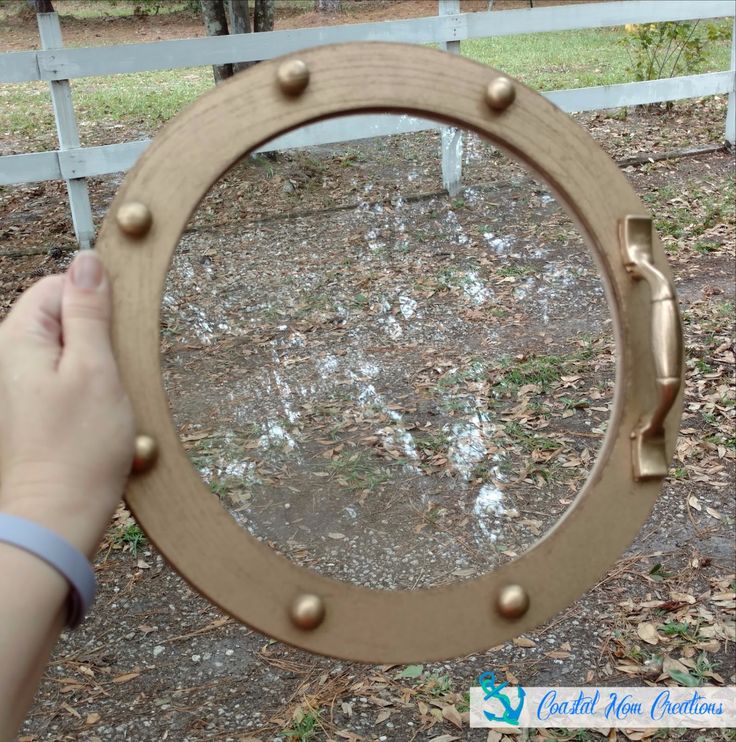 a person holding up a mirror in front of a fence with trees and grass behind it