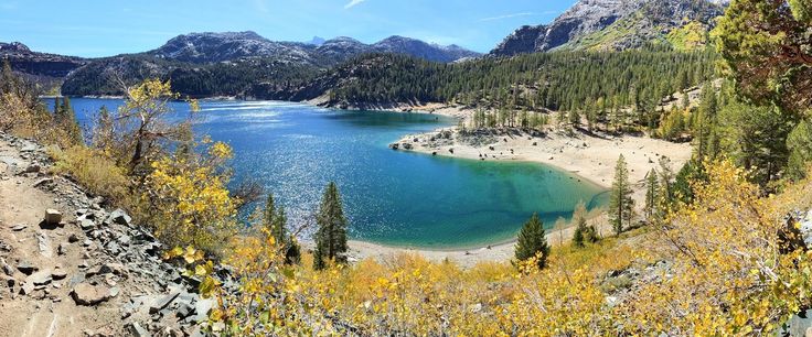 a lake surrounded by mountains and trees with blue water in the foreground on a sunny day