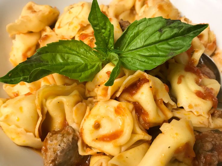 a white bowl filled with pasta and meat covered in sauce, garnished with a green leaf