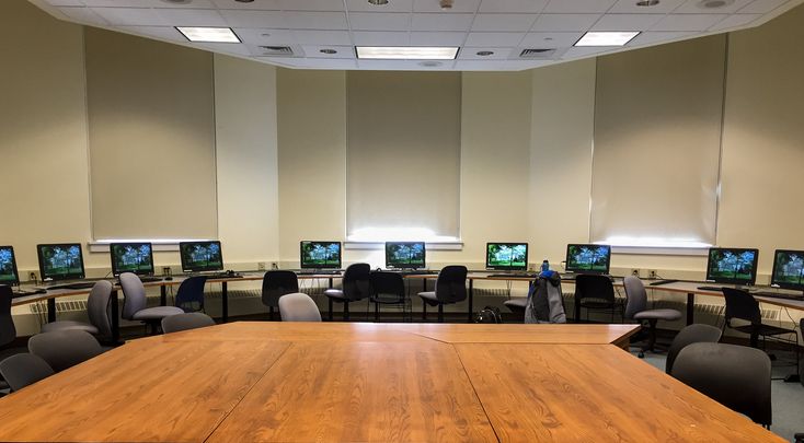 an empty room with many computers on the desks and in front of them is a long wooden table