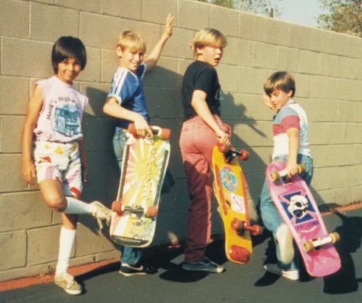 three boys are holding skateboards in front of a brick wall and one boy is pointing at the camera