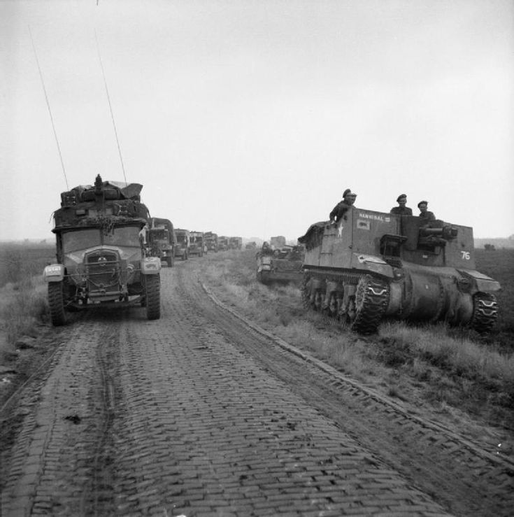 several tanks driving down a dirt road next to grass and trees in the distance with men on top of them