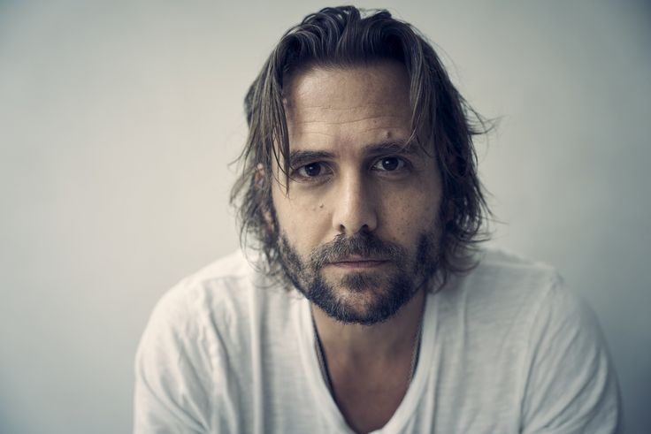 a man with long hair and beard wearing a white t - shirt looking at the camera