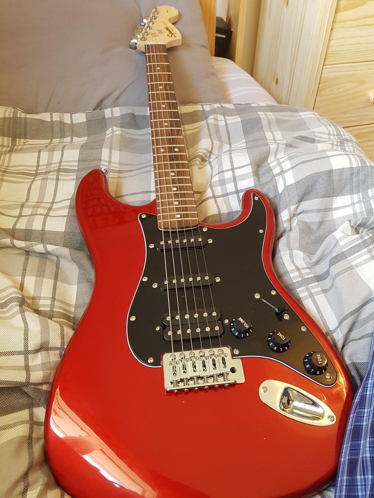 a red electric guitar laying on top of a bed next to a blue and white checkered pillow