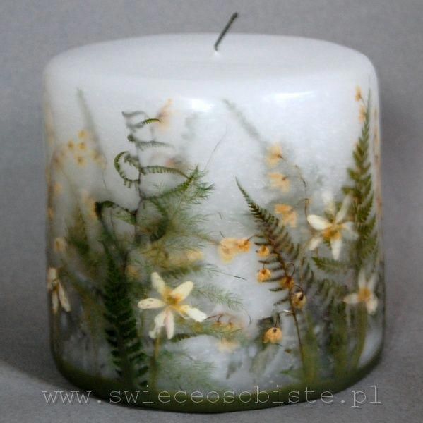 a white candle with yellow flowers and ferns on the inside is sitting in front of a gray background