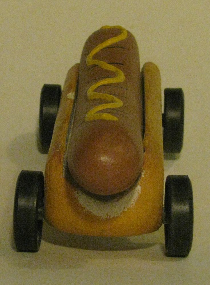 a hot dog in a bun sitting on top of a wooden toy car with wheels