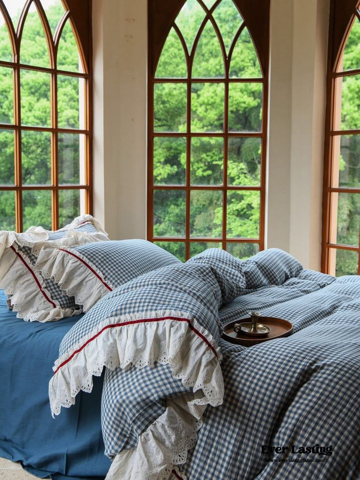 a bed with blue sheets and pillows in front of two large arched windows that look out onto the woods