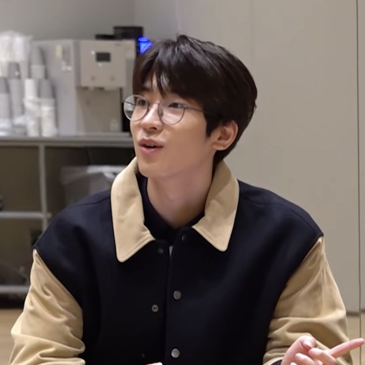 a young man wearing glasses sitting at a table in front of a laptop computer and talking to someone