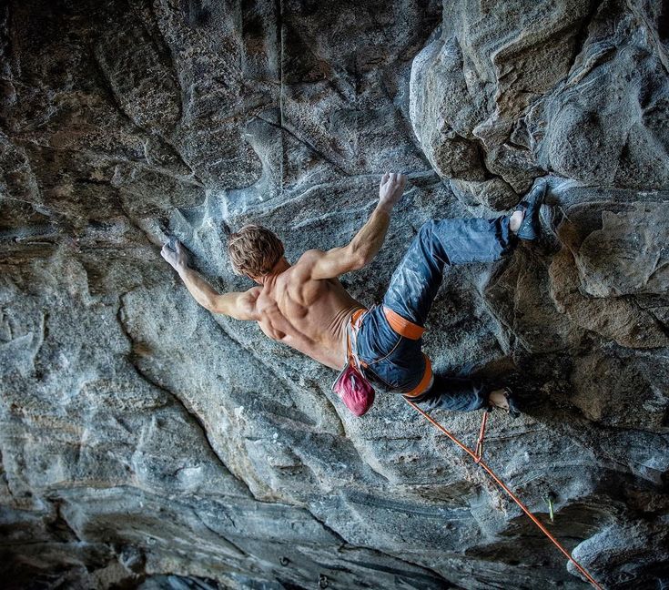 a shirtless man climbing up the side of a rock face with his hands in the air