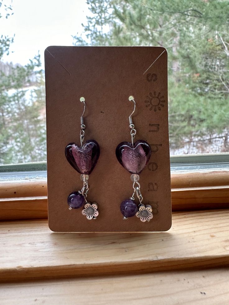 a pair of purple heart shaped earrings sitting on top of a wooden table next to a window