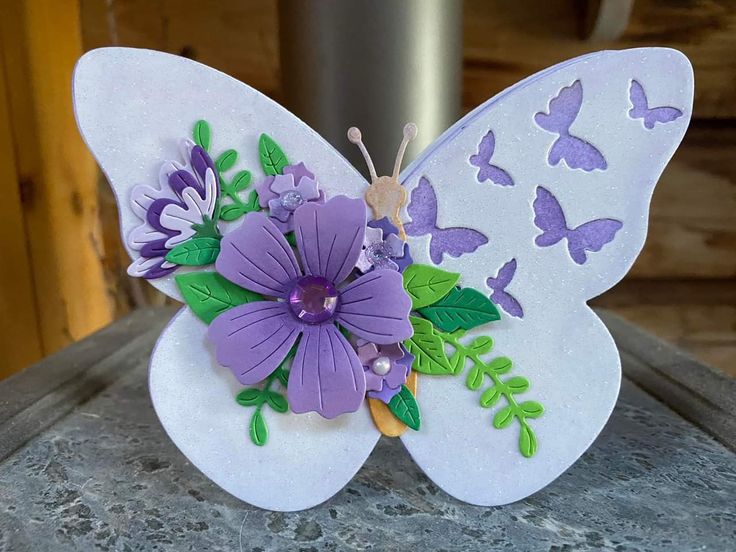 a white butterfly with purple flowers and green leaves on it's wings, sitting on a stone surface