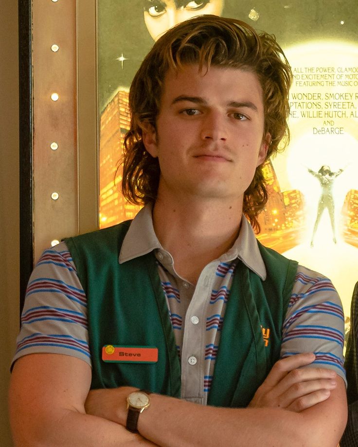 a man with his arms crossed standing in front of a movie poster wearing a green vest