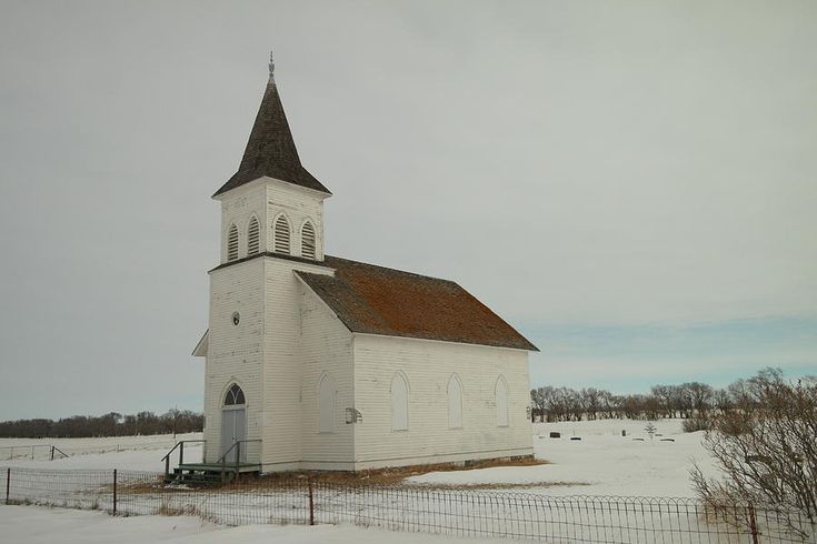 an old white church sits in the middle of a snow covered field with a fence around it