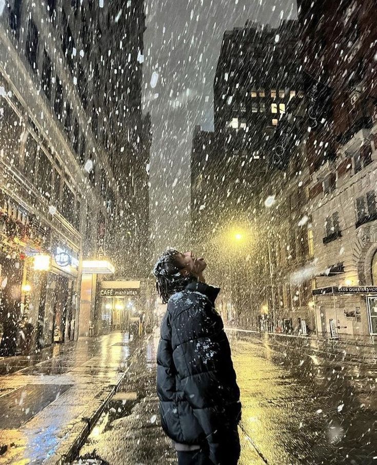 a person standing in the middle of a city street on a rainy day with snow falling all around