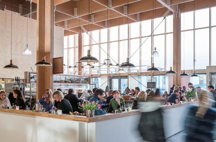 people sitting at tables in a restaurant with lots of windows and hanging lights above them