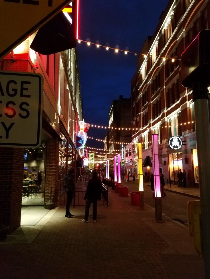 two people are standing on the sidewalk in front of some buildings with lights strung over them