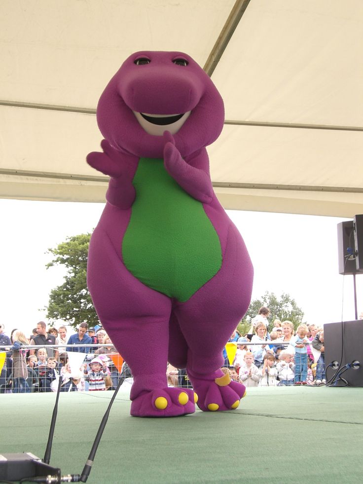 an inflatable purple dinosaur standing on top of a green field next to a crowd