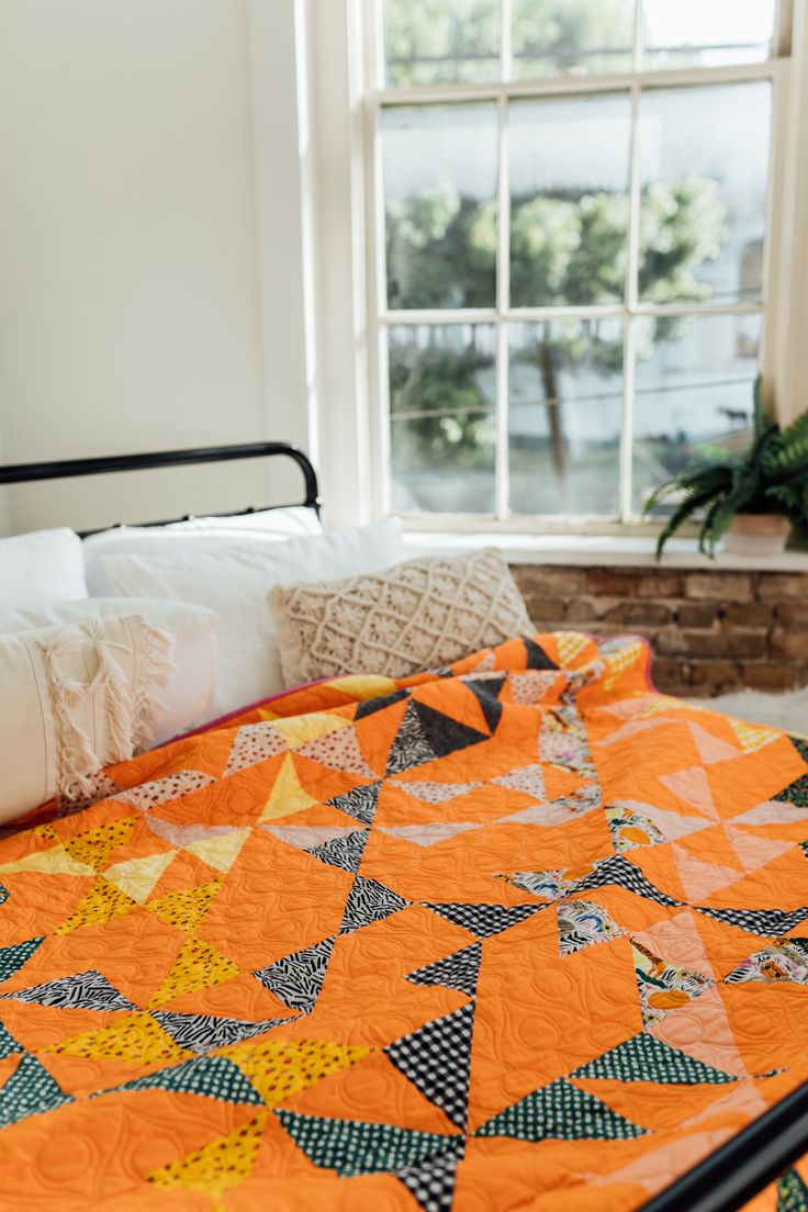 an orange and black quilt on a bed in front of a window with white pillows