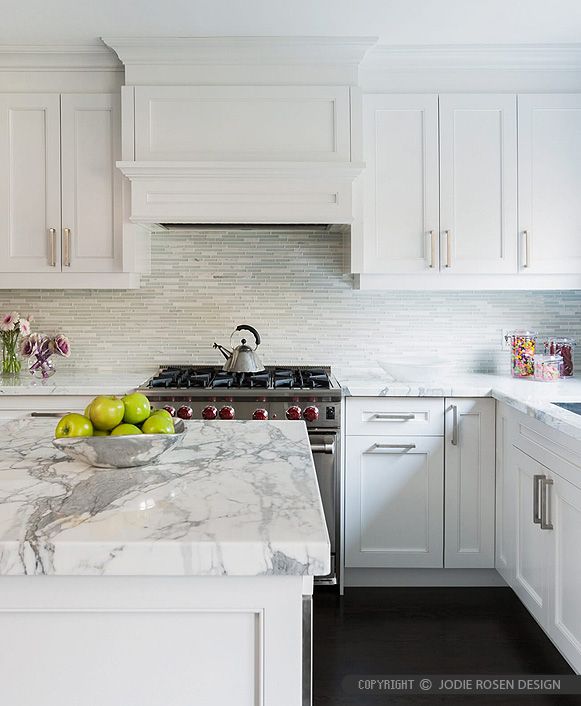 a white kitchen with marble counter tops and stainless steel appliances, along with two green apples in a bowl on the island