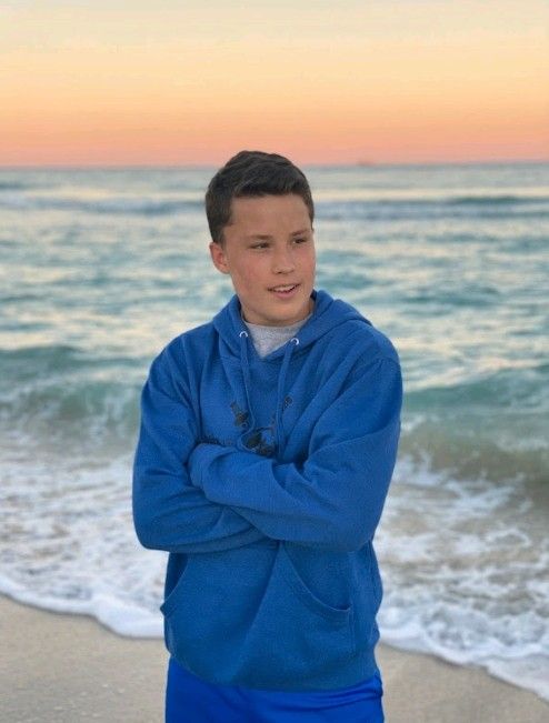 a young boy standing on the beach with his arms crossed and looking at the camera