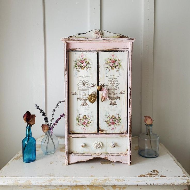 an old white cabinet with flowers and vases on the side table next to it