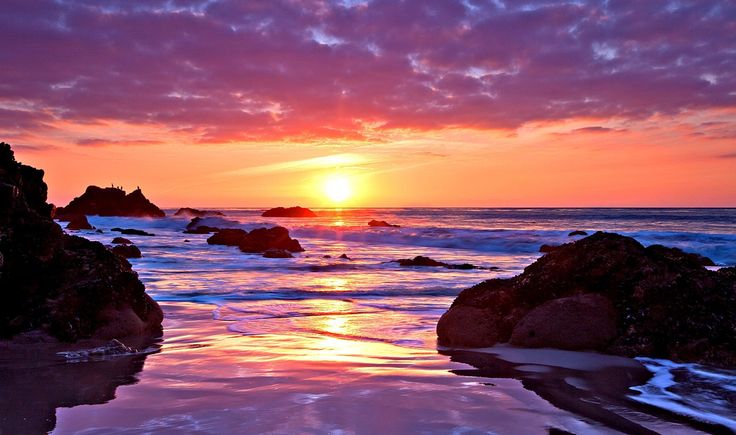 the sun is setting over the ocean with rocks in the foreground and water on the ground