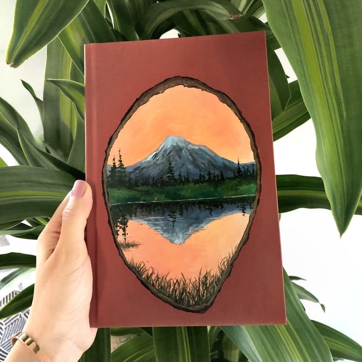 a person holding up a book in front of a plant with a mountain scene on it