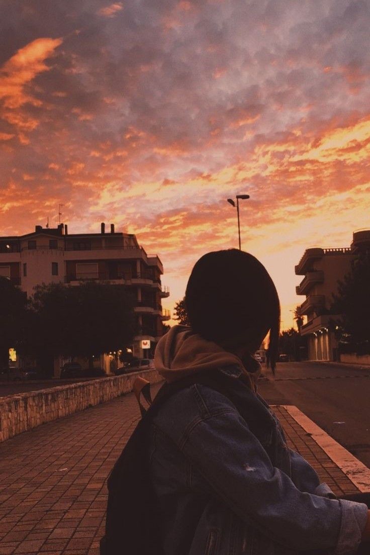 a person sitting on a bench looking at the sky with buildings in the background as the sun sets