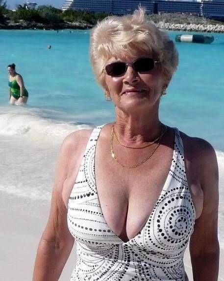 an older woman in a white swimsuit standing on the beach with her hands in her pockets