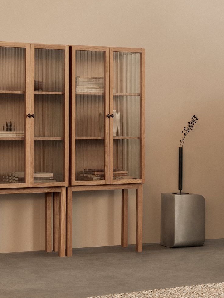 a tall wooden cabinet with glass doors next to a vase on the floor in front of a beige wall