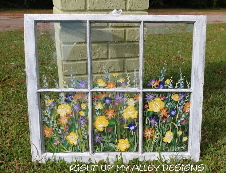 an old window is painted with flowers and grass in front of a brick wall that reads right up my alley designs