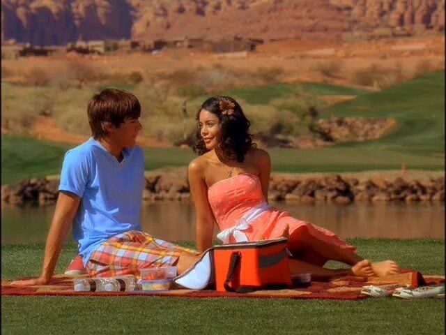 a man and woman sitting on a blanket next to a body of water with mountains in the background