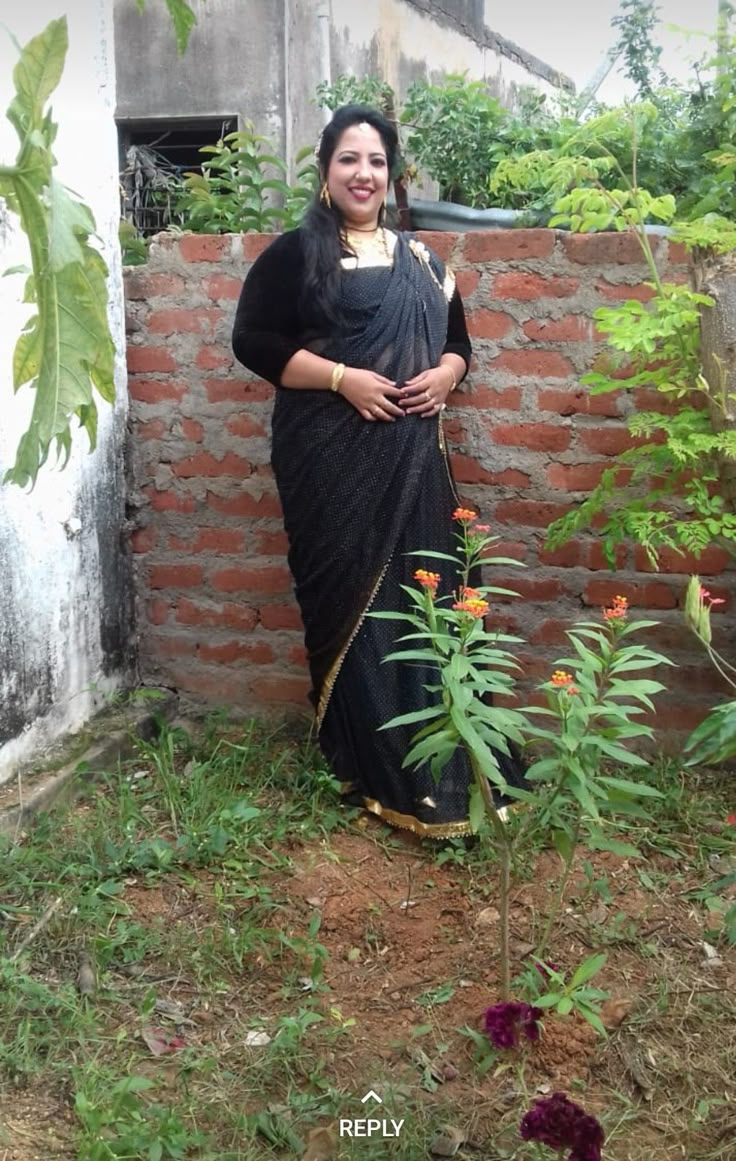 a woman standing in front of a brick wall next to a small tree and flowers