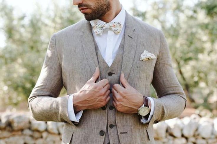 a man in a suit and bow tie standing next to some trees with his hands on his chest