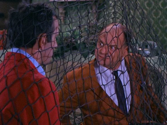 two men in orange jackets and ties looking at each other through a fenced area