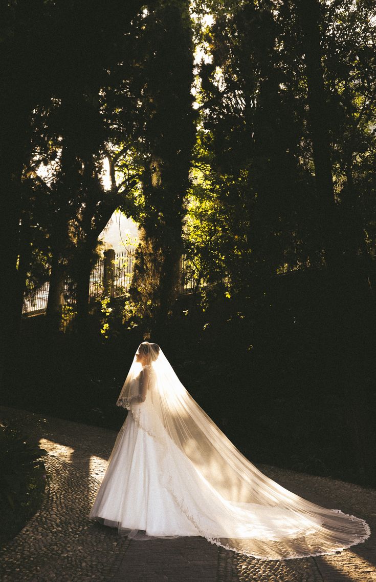 a woman in a wedding dress and veil is walking through the park with her back to the camera