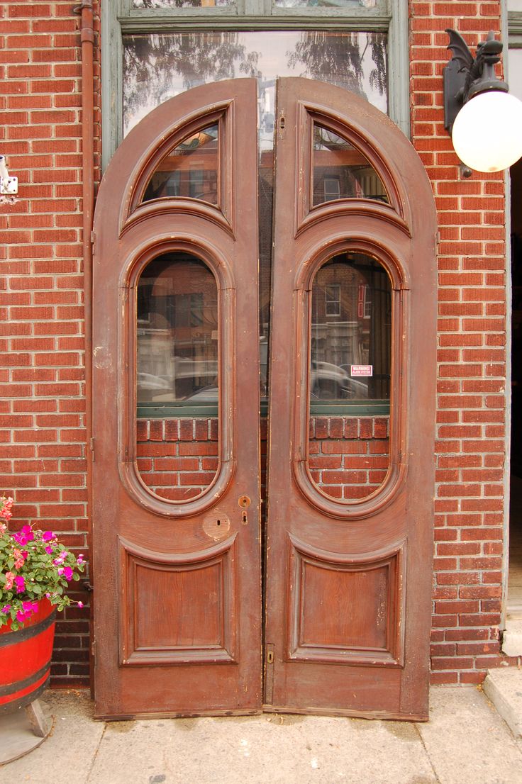 two wooden doors are open in front of a brick building with potted flowers on the sidewalk