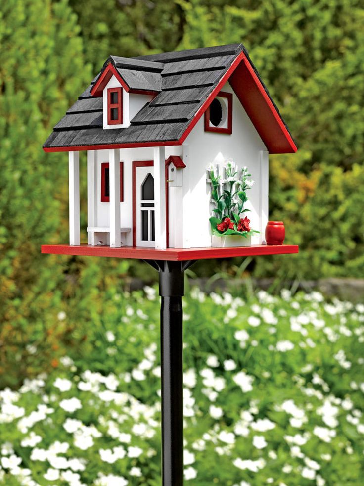 a red and white bird house on top of a black pole in front of flowers