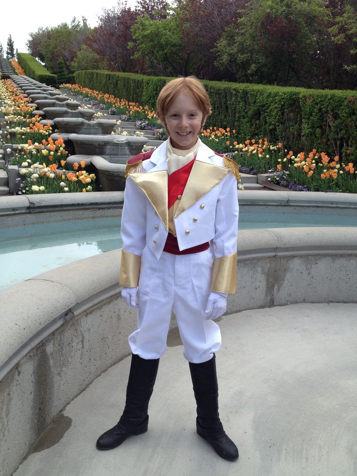 a young boy dressed in a white and gold costume standing next to a fountain with orange flowers