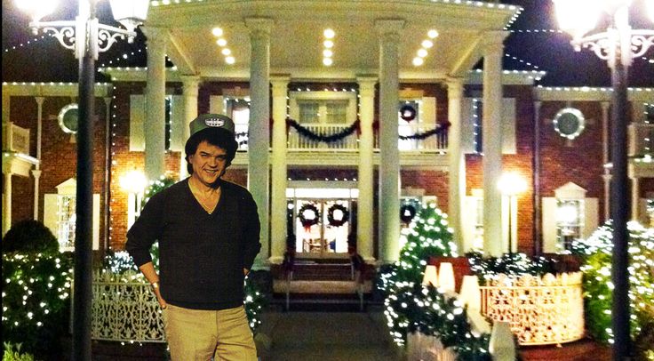 a man is standing in front of a house with christmas lights on the porch and trees