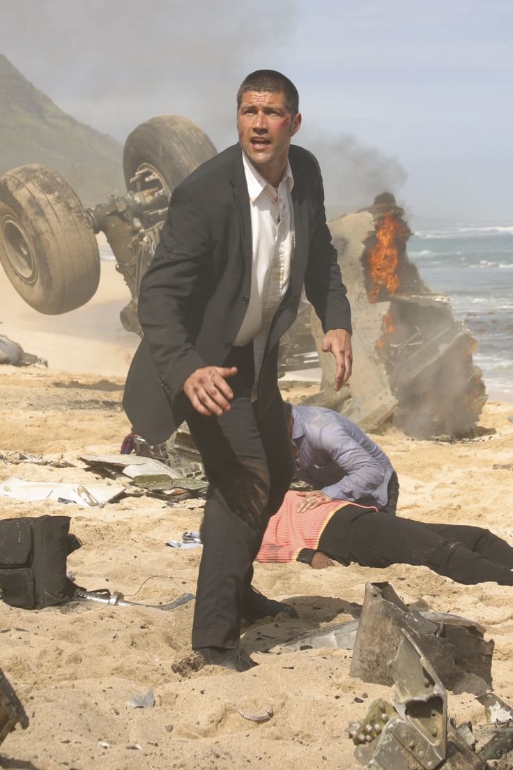 a man in a suit and tie standing next to another man on the beach near an overturned car