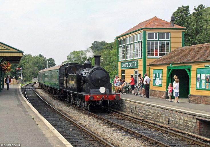 a black train traveling past a train station next to a green and yellow building with people standing on the platform