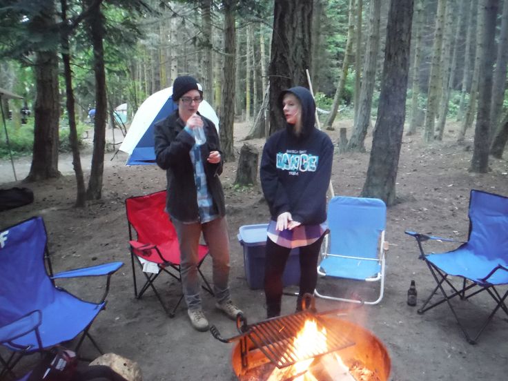 two women standing next to each other in front of a campfire with chairs and umbrellas