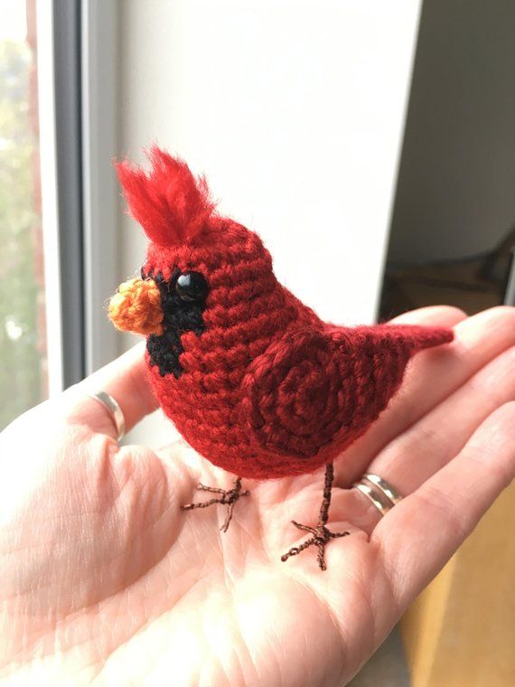 a small red crocheted bird sitting on someone's hand in front of a window