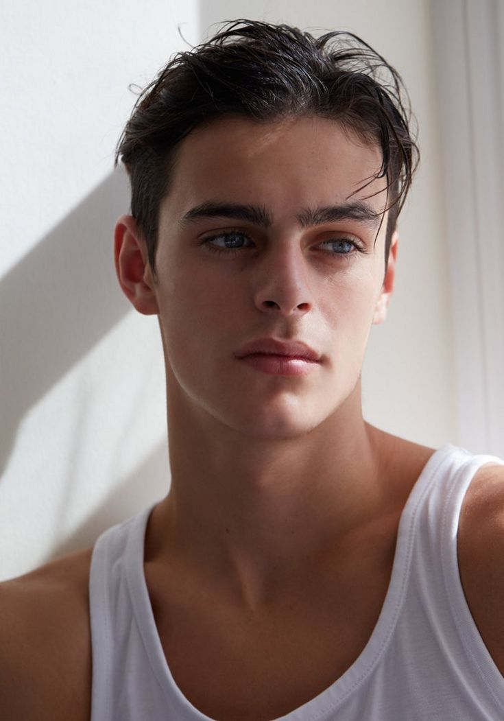 a young man wearing a white tank top looking at the camera while standing in front of a window