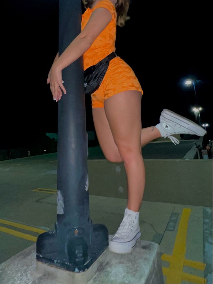 a woman in an orange outfit leaning on a pole with her legs spread out and one leg bent