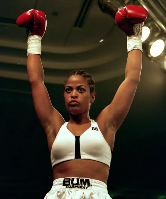 a female professional boxer raises her arms in the air while wearing red boxing gloves and white shorts