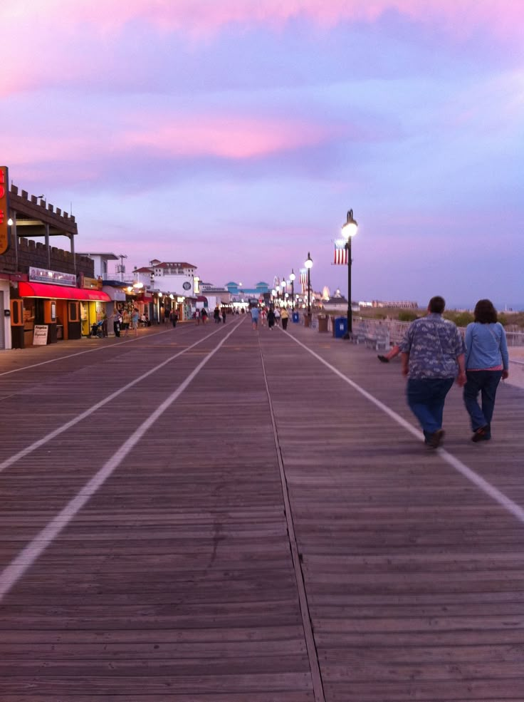 two people are walking down the boardwalk towards some shops and restaurants at dusk or dawn