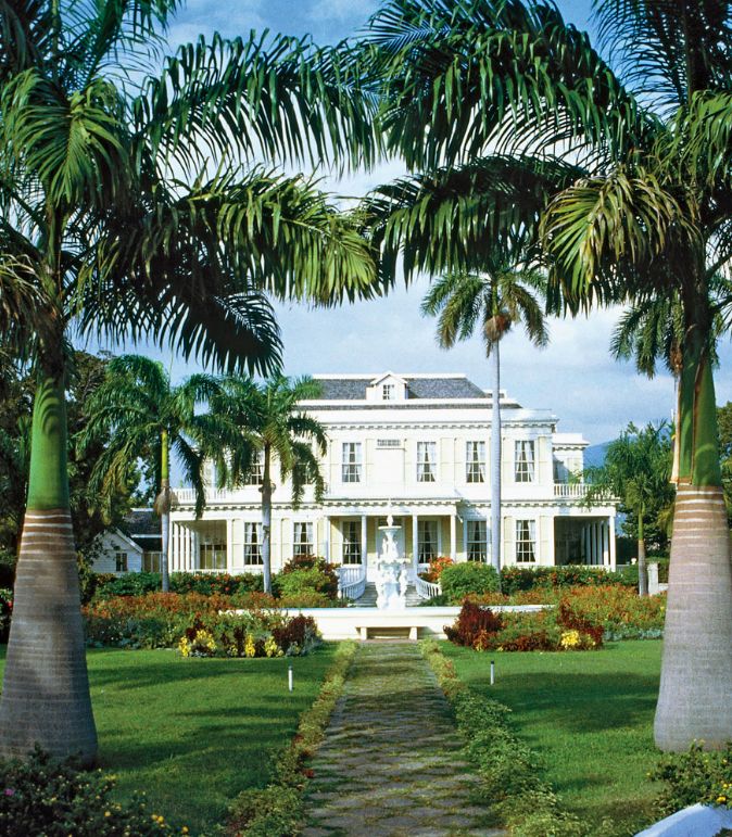 palm trees in front of a large white house with a fountain surrounded by greenery